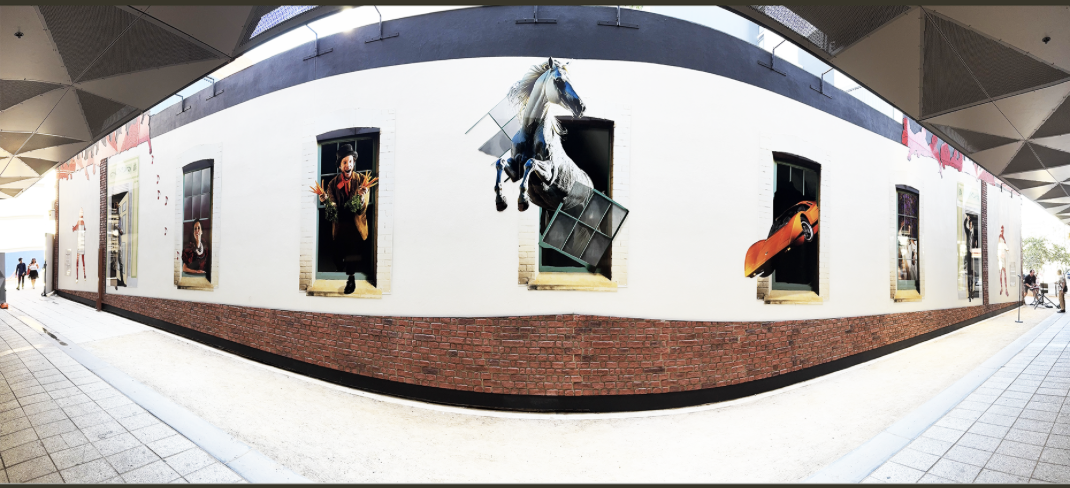 The wall photographed as a panorama: white render over a red brick base, under a canopy, hung with printed panels of figures standing in doorways and windows, a white horse rearing out through a broken window frame and an orange sports car nosing out of another.