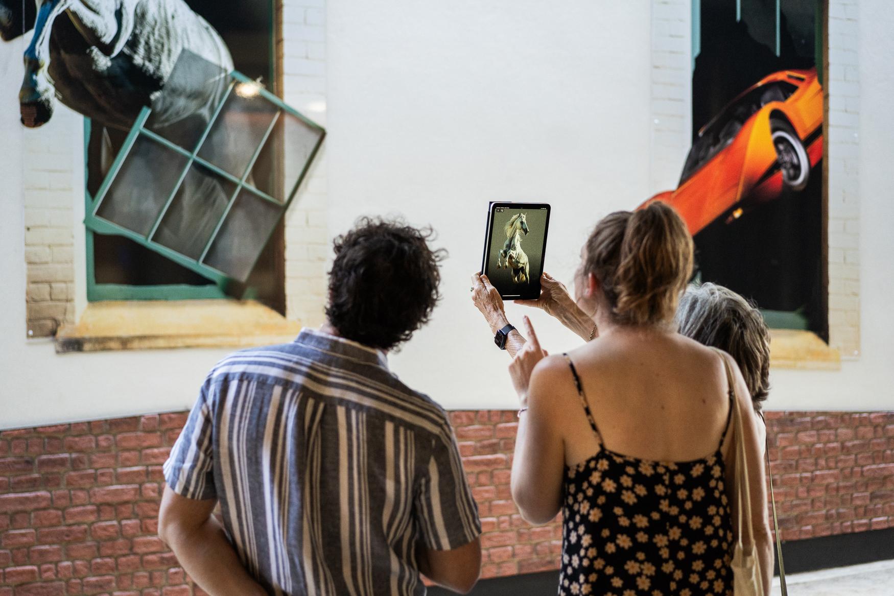 Three people stand at the wall with their backs to the camera; one holds up a tablet, and on its screen the horse painted on the wall behind is shown mid-stride.