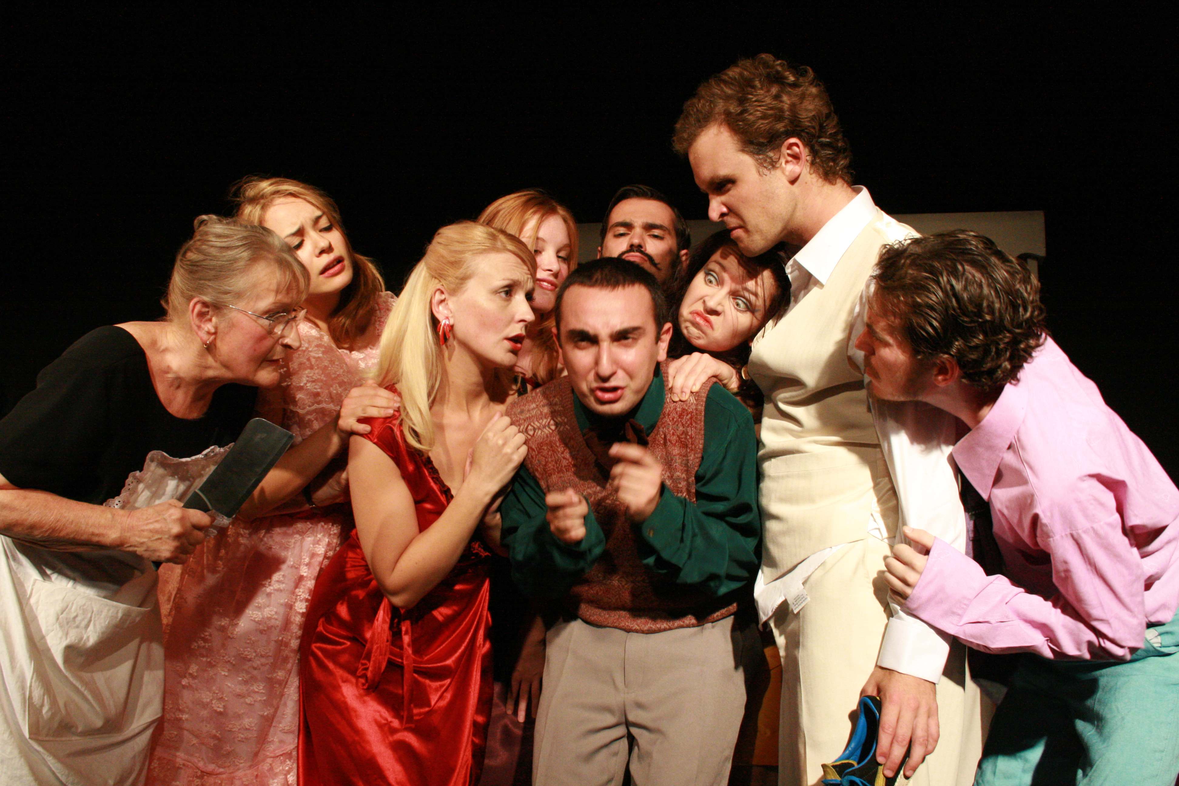 The full cast crowd in around a cowering Myles Fletcher on a black stage — an older woman brandishing a meat cleaver at the left edge, the rest in evening dress, every face suspicious.