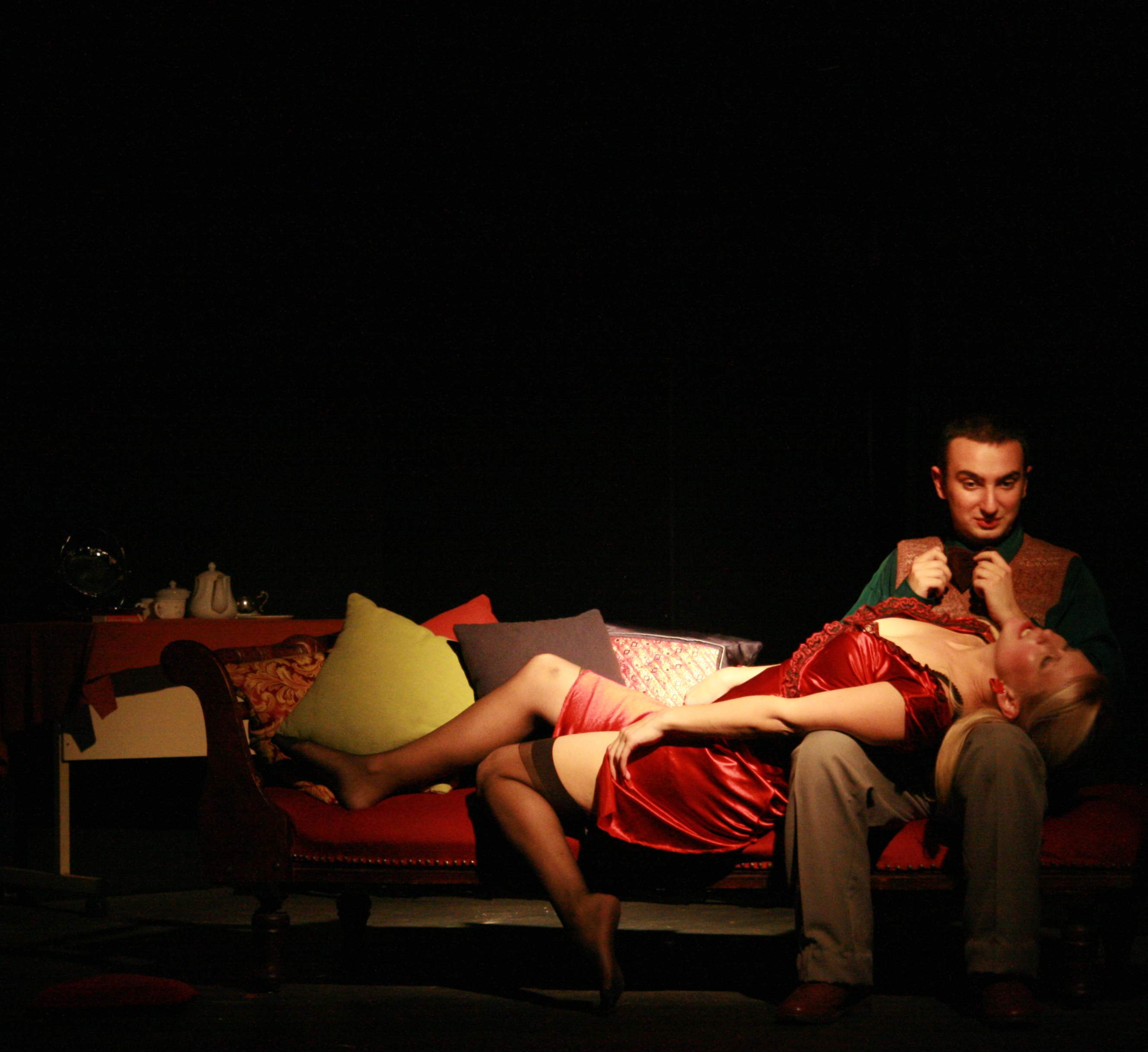 A woman in a red satin dress lies back across a chaise longue while a man in a green shirt and knitted vest adjusts his bow tie above her, lit in a single warm pool against the dark.