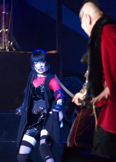 A performer in white face paint and heavy black eye make-up, in a black cape over red and black vinyl, stands in profile beside a figure in a red coat.