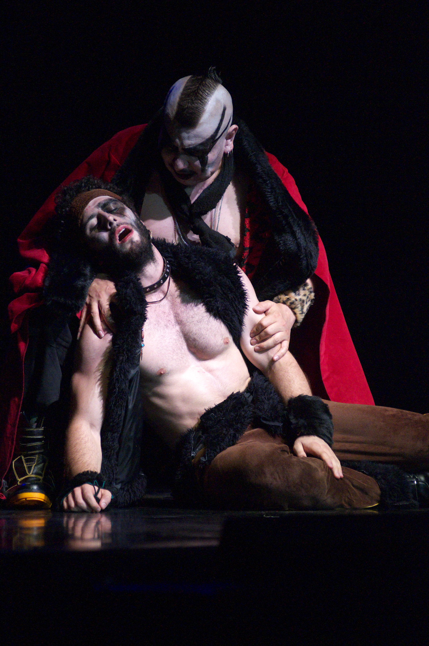 A figure in a red cloak and mohawk, face painted white and streaked with black, holds a bearded performer in a fur-trimmed coat as he falls back across the stage floor.