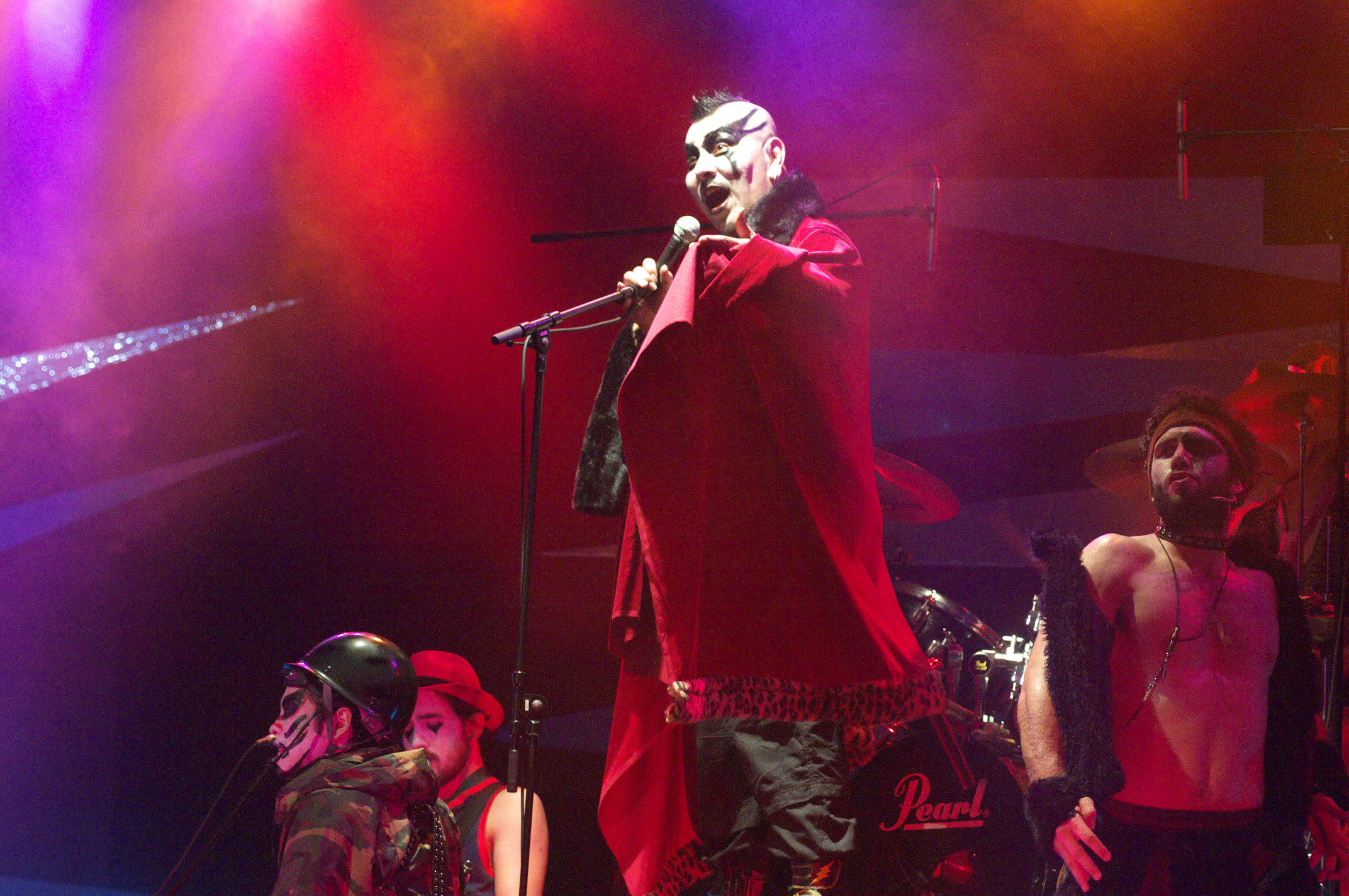 A singer in a long red coat and white face paint takes the microphone under red and purple stage lights, the band and company arrayed behind him around a drum kit.