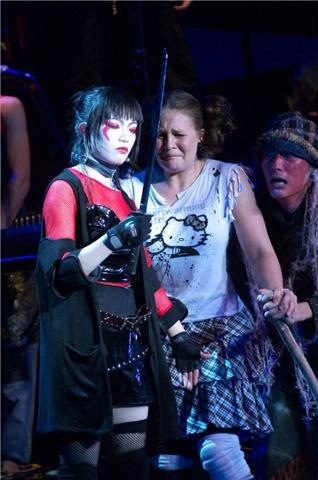 A performer in white face paint and a red-and-black costume stands beside a schoolgirl in a Hello Kitty T-shirt, mid-cry, with another figure watching from the dark behind them.