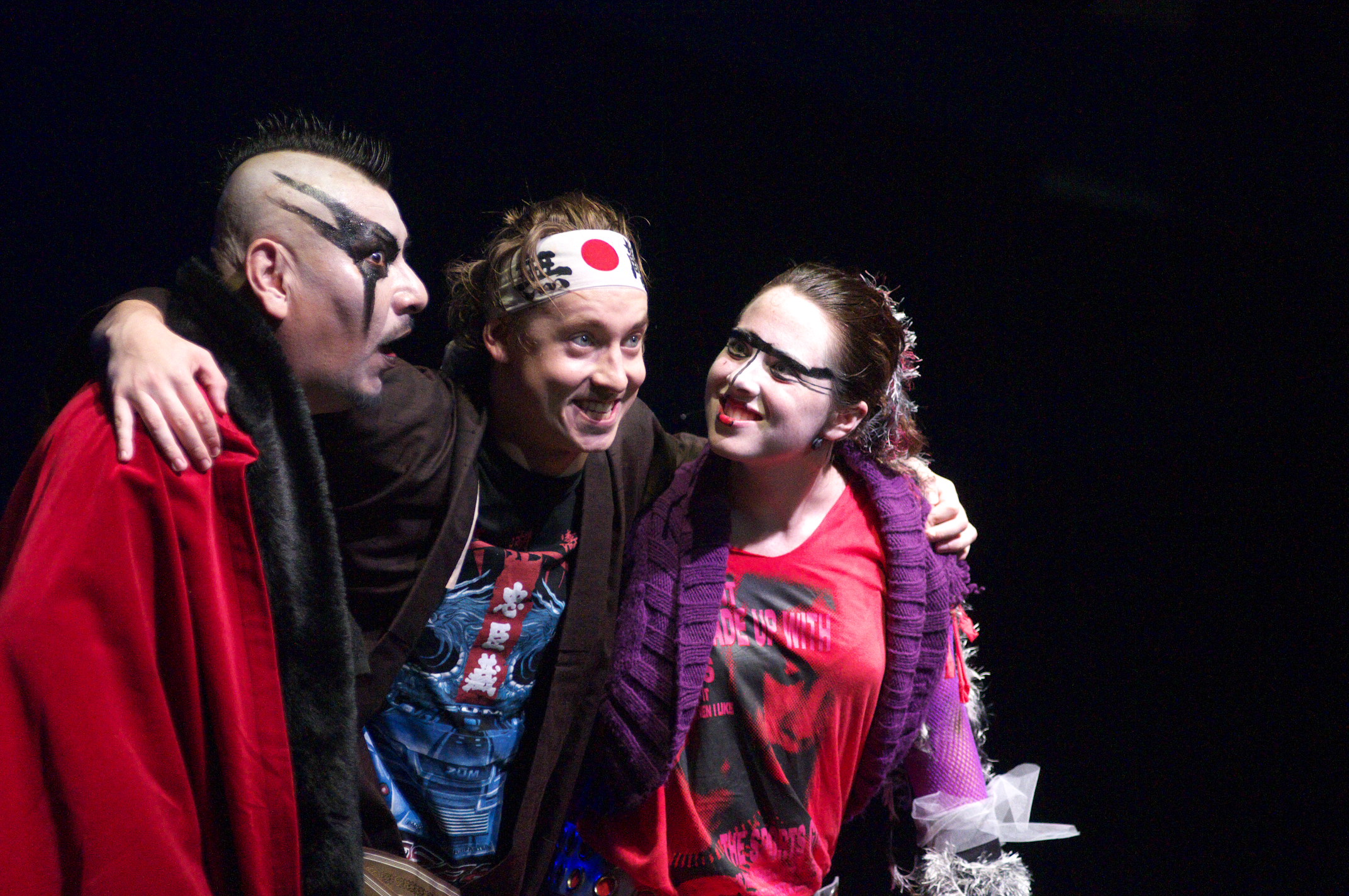 Three of the company with their arms around one another, laughing — a figure in a red cloak and white face paint, a young man in a rising-sun headband, and a woman in a purple scarf.