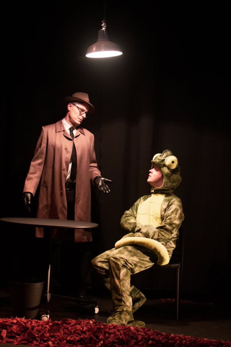 A trench-coated, fedora-wearing interrogator looms over an activist in a lizard costume beneath a single hanging lamp.