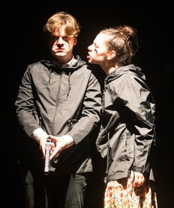 Two actors in hooded jackets stand close together under a hard side light, one turning to speak to the other.