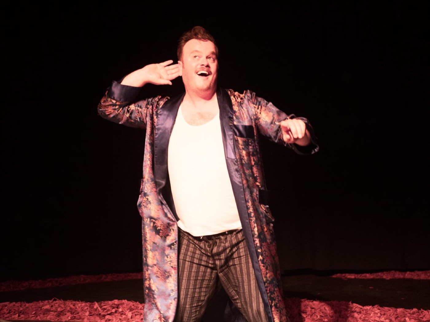 An actor in a patterned dressing gown mimes a gun to his own head, mid-laugh, on a dark stage.